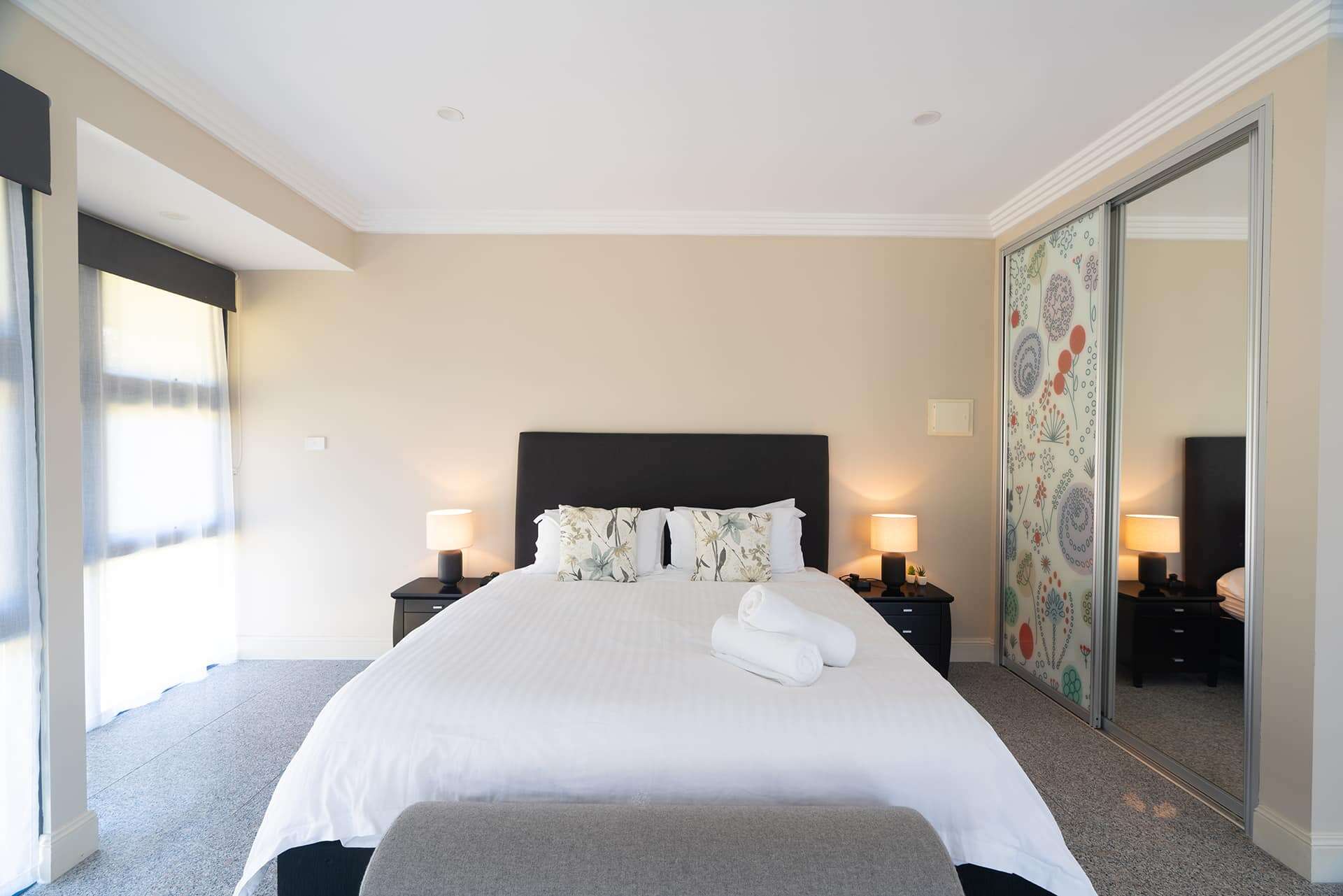 Home 3 Bright contemporary bedroom featuring king-sized bed, stylish lamps, decorative pillows, and large mirrored closet.